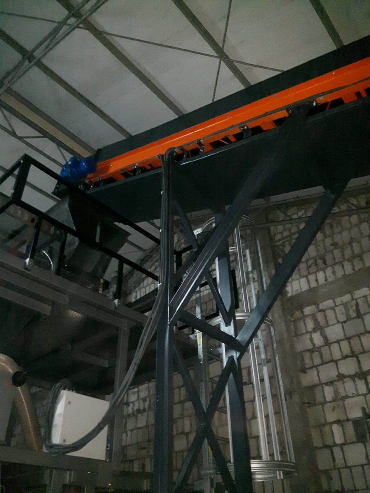 Wnętrze hali produkcyjnej z widoczną konstrukcją stalową podtrzymującą pomarańczowo-czarny system transportowy; szara ściana z cegieł w tle.