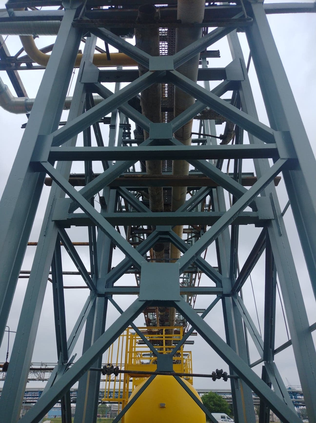 Skomplikowana konstrukcja stalowa z licznymi rurami i drabinkami, widok z dołu, szaro-żółta kolorystyka.