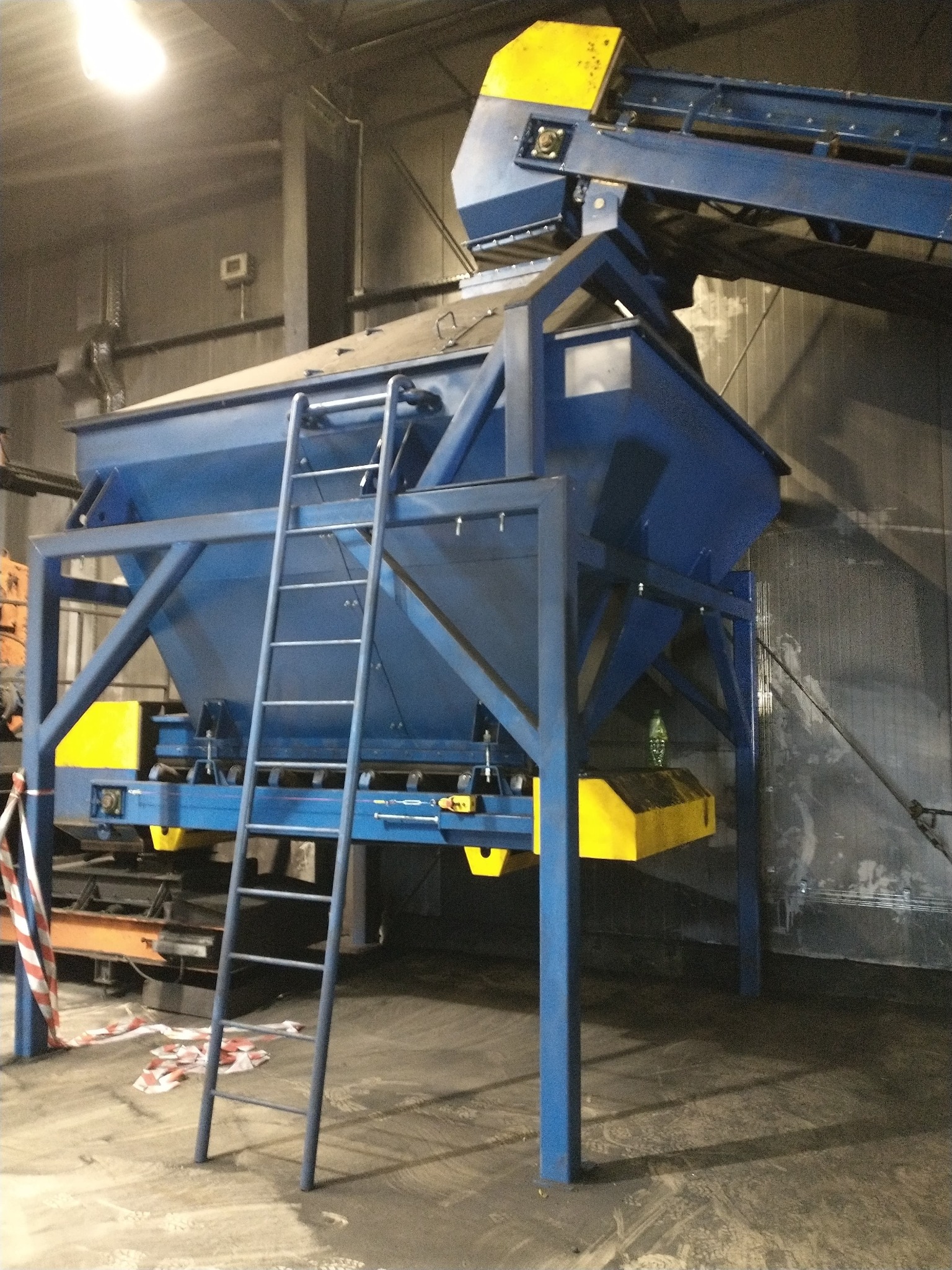 Niebieska konstrukcja stalowa z żółtymi elementami, prawdopodobnie część linii produkcyjnej, z drabinką, widoczna w hali przemysłowej.