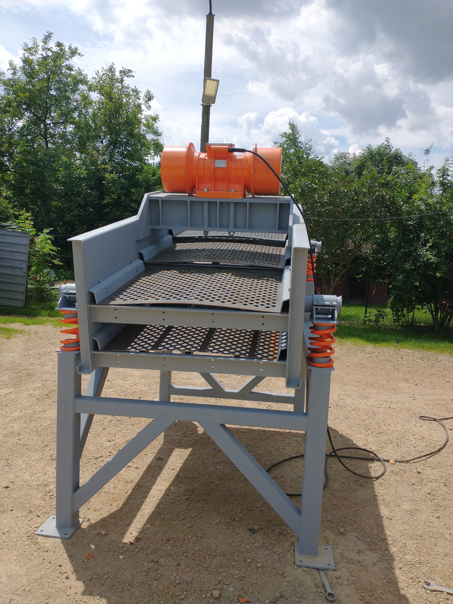 Stacjonarna przesiewacz wibracyjny z pomarańczowym silnikiem, szarą konstrukcją i sprężynami, ustawiony na żwirowym podłożu w otoczeniu zieleni.
