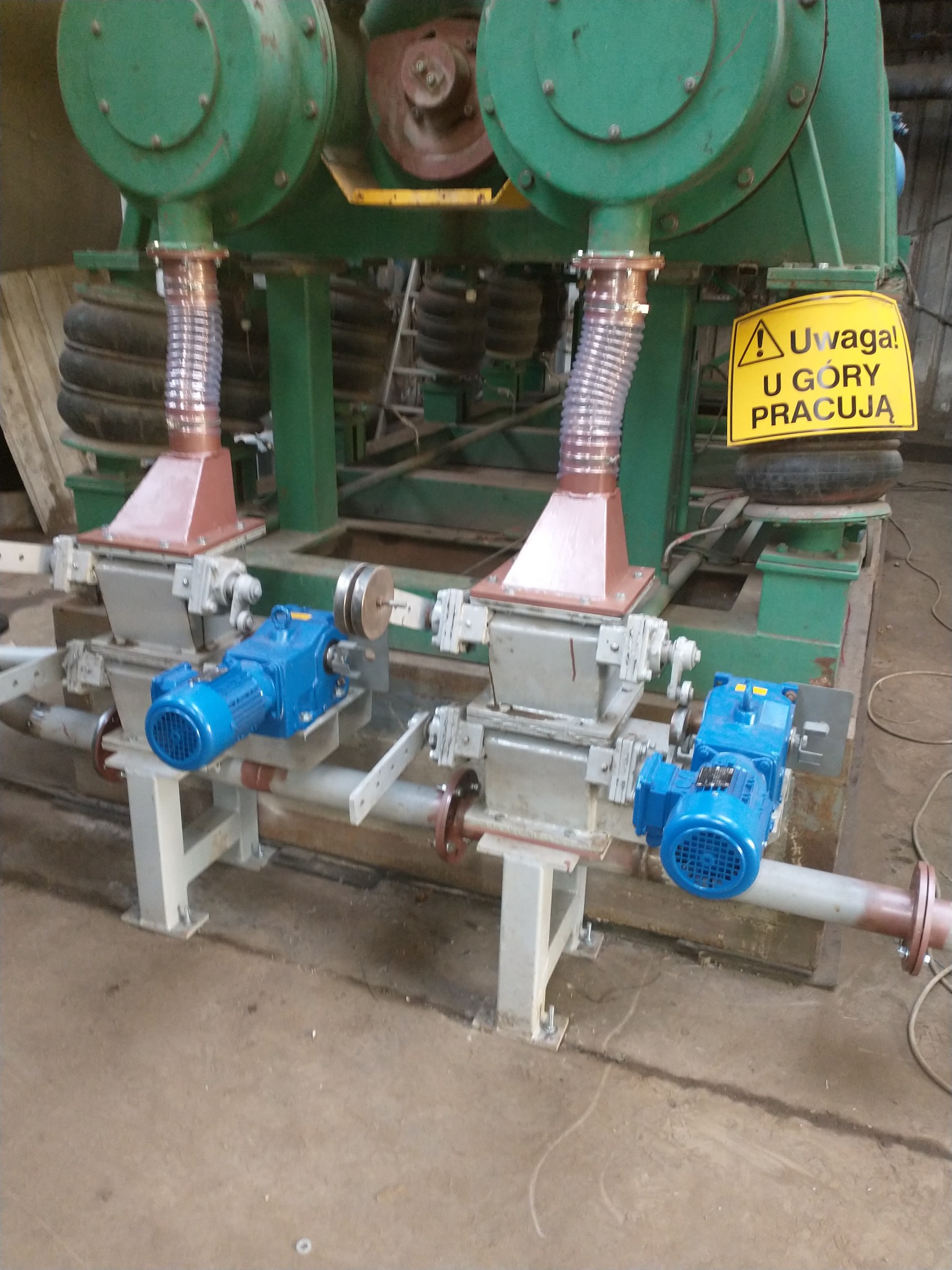 Fragment konstrukcji hali przemysłowej z dwoma silnikami elektrycznymi w kolorze niebieskim, podłączonymi do mechanizmu. Widoczna tabliczka ostrzegawcza 'Uwaga! U góry pracują'.