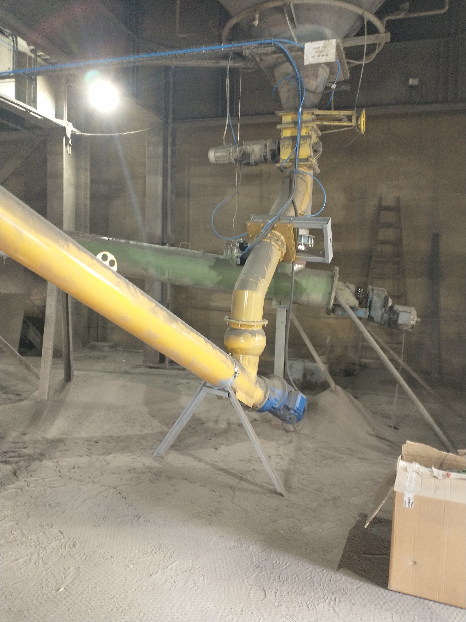 Żółty rurociąg przemysłowy z elementami mocującymi i silnikiem elektrycznym, umieszczony w hali produkcyjnej z betonowymi ścianami i podłożem pokrytym pyłem.