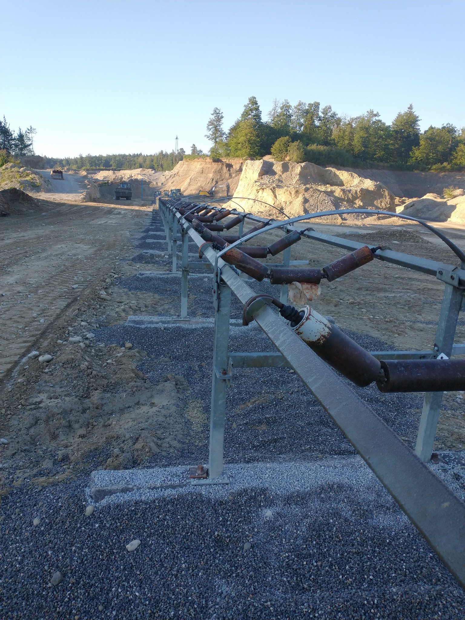 Długa, stalowa konstrukcja przenośnika taśmowego z rdzewiejącymi rolkami w kamieniołomie, z koparkami i ciężarówkami w tle.