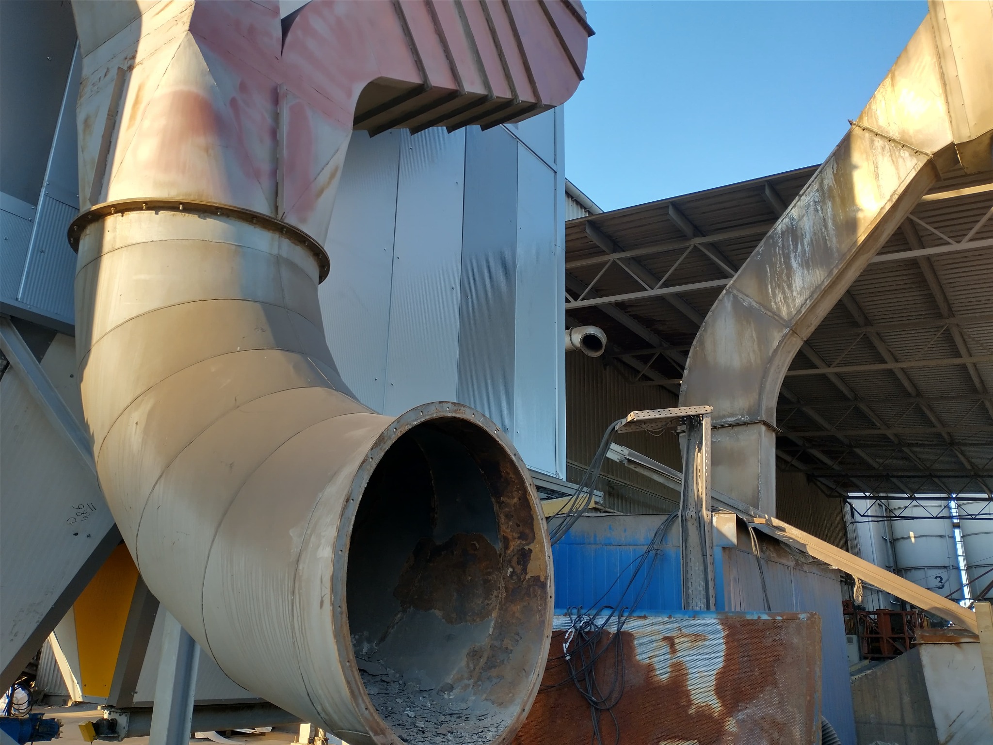 Zewnętrzny system wentylacyjny z rur stalowych o dużym przekroju, z widocznymi śladami korozji i zabrudzeń, zamontowany przy hali przemysłowej z blachy trapezowej.