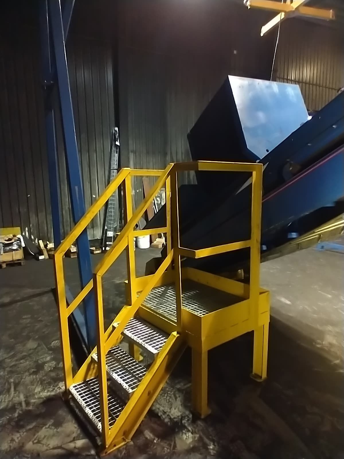 Żółte metalowe schody z poręczami prowadzące do podestu w hali przemysłowej, widoczne elementy konstrukcji i podajnika.