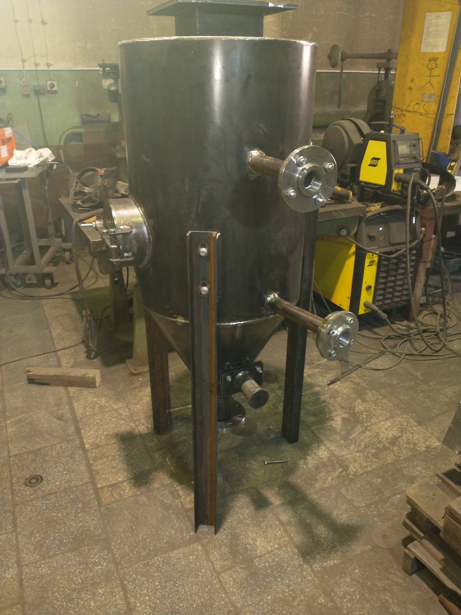 Cylindryczny zbiornik metalowy z przyspawanymi rurami i kołnierzami, wsparty na stalowych nogach, widoczny w warsztacie z różnymi urządzeniami do obróbki metali w tle.