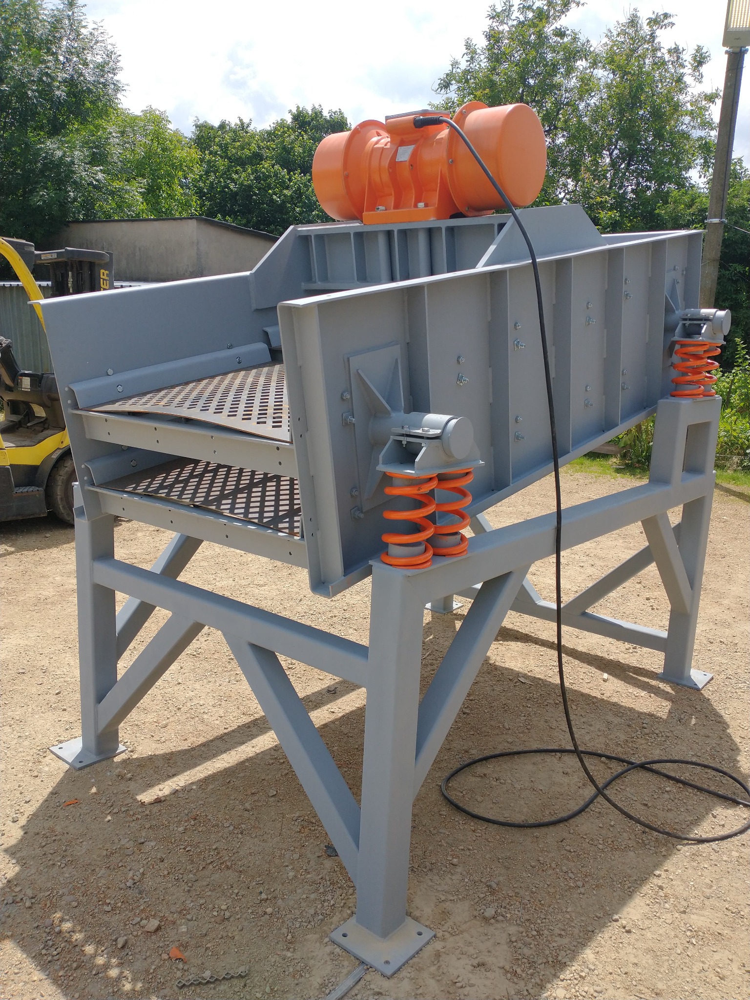 Szara, metalowa konstrukcja przesiewacza wibracyjnego z pomarańczowym silnikiem wibracyjnym na górze i sprężynami amortyzującymi, stojąca na żwirowym podłożu, z widocznym w tle wózkiem widłowym.