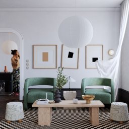 Umbra Studio - Jasne wnętrze z zielonymi fotelami, drewnianym stolikiem kawowym i geometrycznym dywanem w szachownicę, uzupełnione przez dekoracje i osobę rozmawiającą przez telefon w tle.