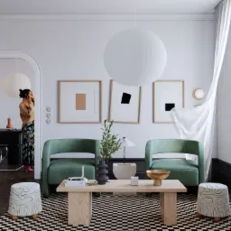 Jasne wnętrze z zielonymi fotelami, drewnianym stolikiem kawowym i geometrycznym dywanem w szachownicę, uzupełnione przez dekoracje i osobę rozmawiającą przez telefon w tle.