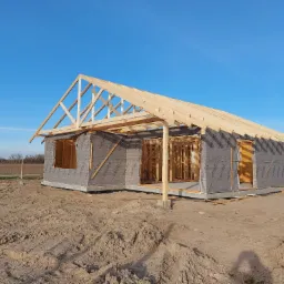 Surowy stan budowy domu: widoczna drewniana konstrukcja szkieletowa z zamontowaną membraną, odsłonięte krokwie dachu, teren budowy z piaskiem i błękitne niebo w tle.