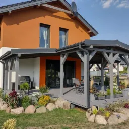 Nowoczesny dom z pomarańczową elewacją i szarą drewnianą pergolą połączoną z tarasem, otoczony ogrodem z kamieniami i roślinami.