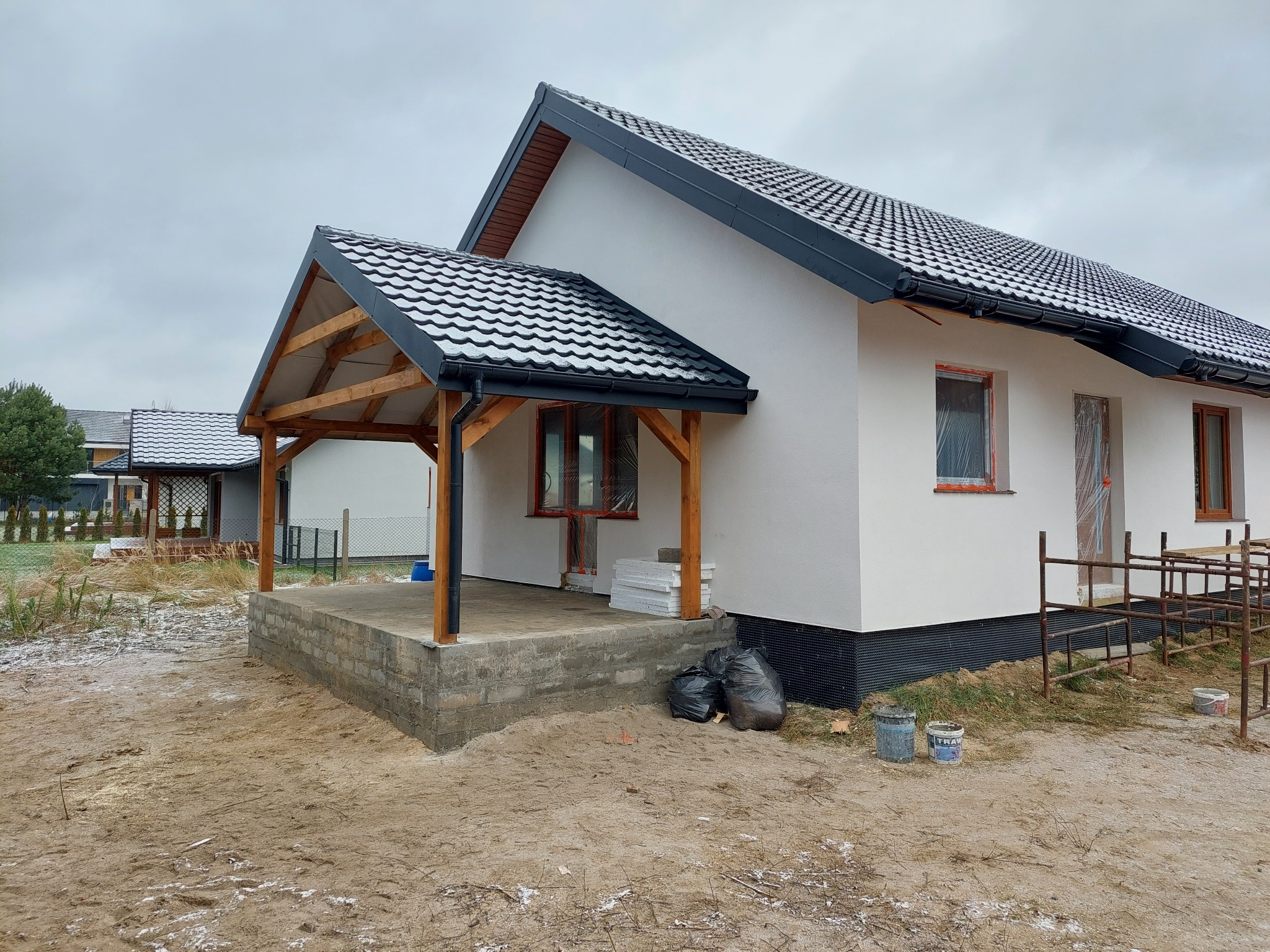 Niedokończony parterowy dom z białymi ścianami, czarnym dachem z dachówki i drewnianą konstrukcją zadaszenia werandy, widoczne materiały budowlane i rusztowania.