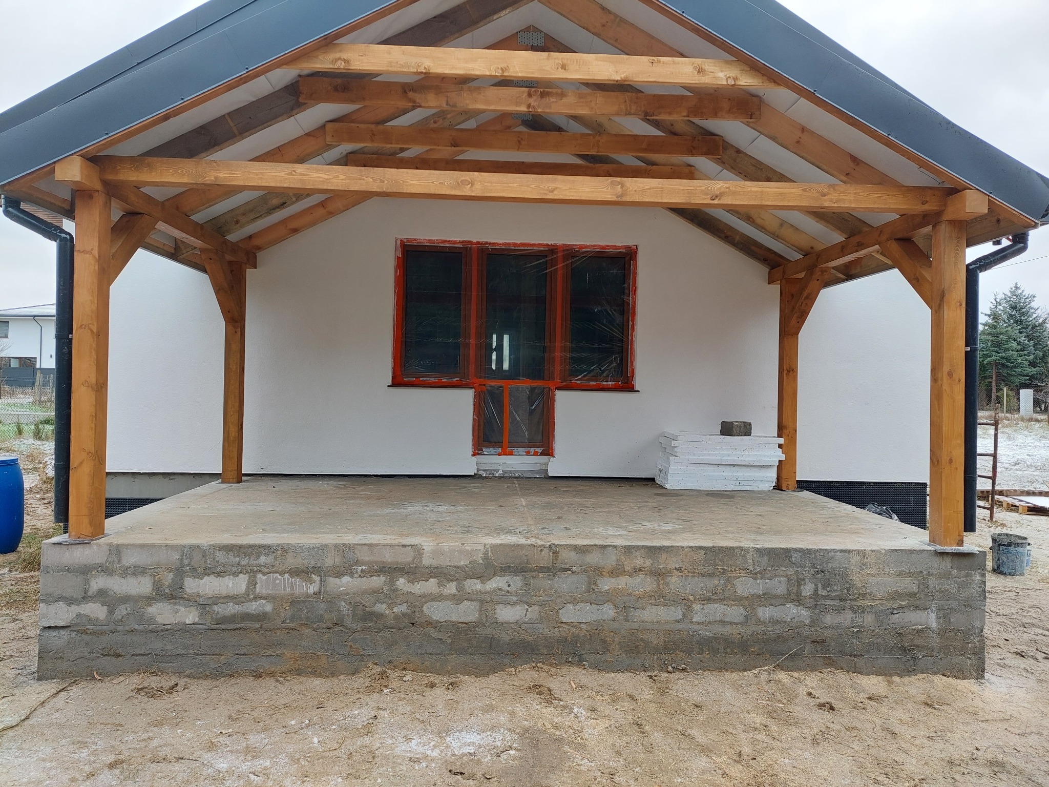 Surowy stan budynku szkieletowego z drewnianą konstrukcją zadaszenia, widoczne okno zabezpieczone folią, niezadaszony taras z bloczków betonowych, materiały budowlane obok.