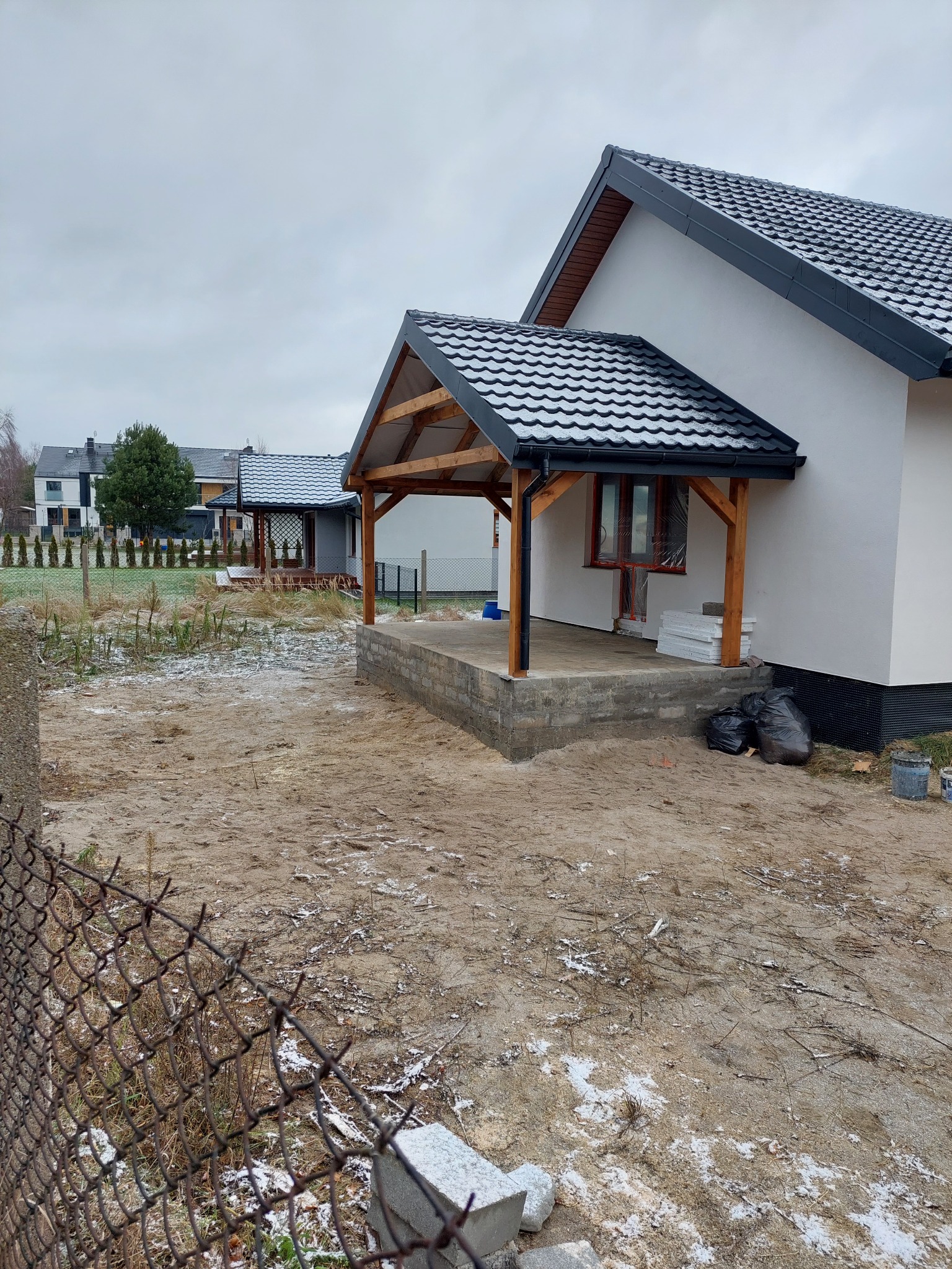Surowy stan budowy domu jednorodzinnego z zadaszonym wejściem, widoczne drewniane słupy i betonowy fundament, w tle drugi dom i ogrodzenie z siatki, ziemia pokryta resztkami śniegu.