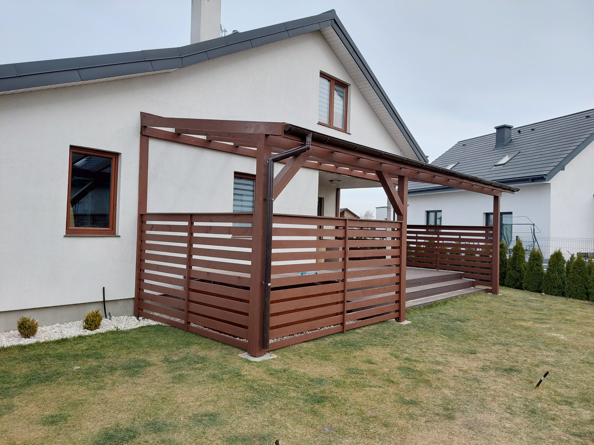 Przydomowa, brązowa konstrukcja z drewna, częściowo zabudowana poziomymi deskami, tworząca zacienioną przestrzeń tarasową przy białym domu z szarym dachem. Widoczne elementy drewnianego tarasu...