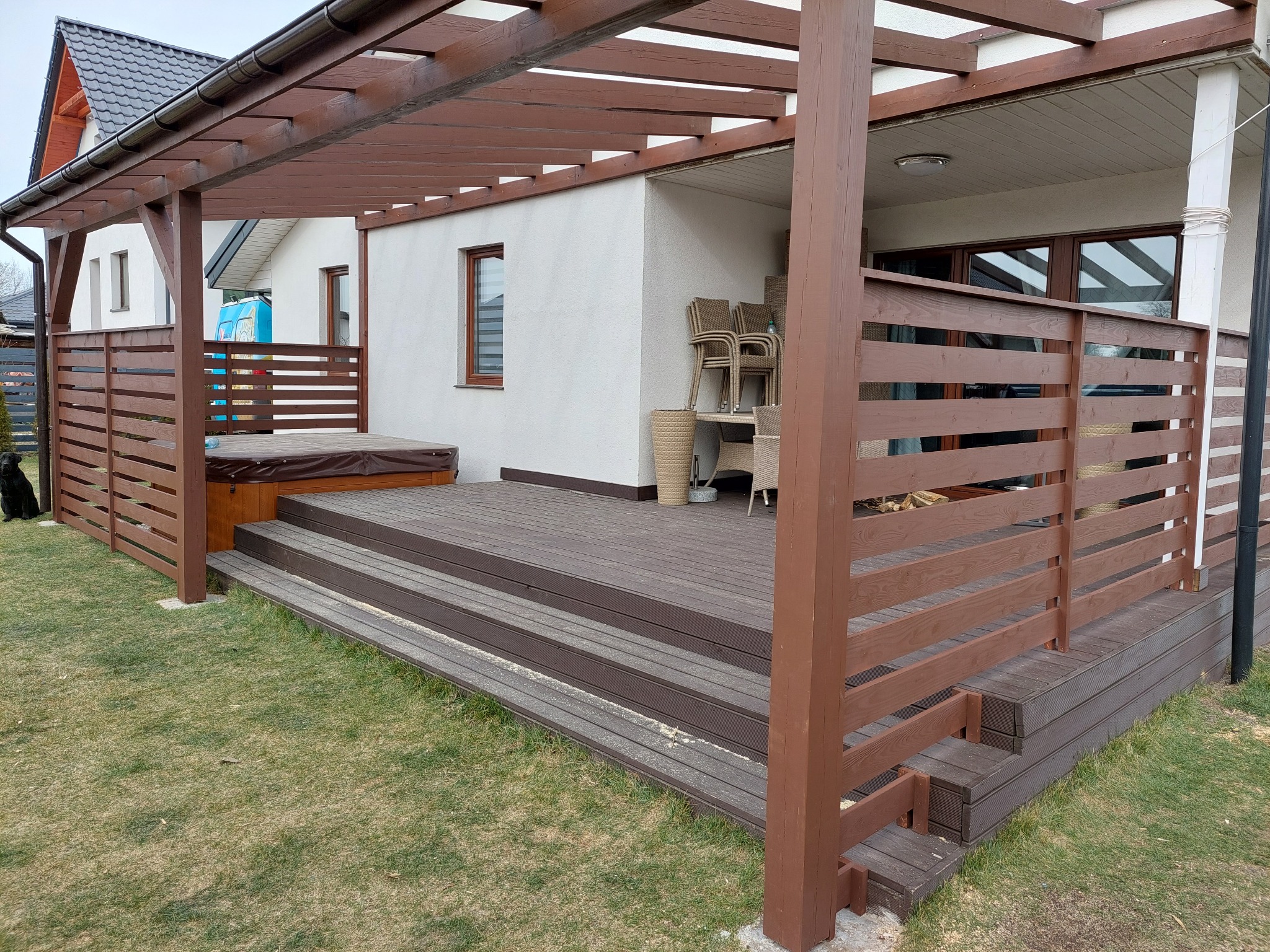 Zadaszony taras z kompozytowych desek w kolorze ciemnego brązu, z drewnianą pergolą w tym samym odcieniu, otoczony zielonym trawnikiem. Widoczna zabudowa jacuzzi i meble ogrodowe.