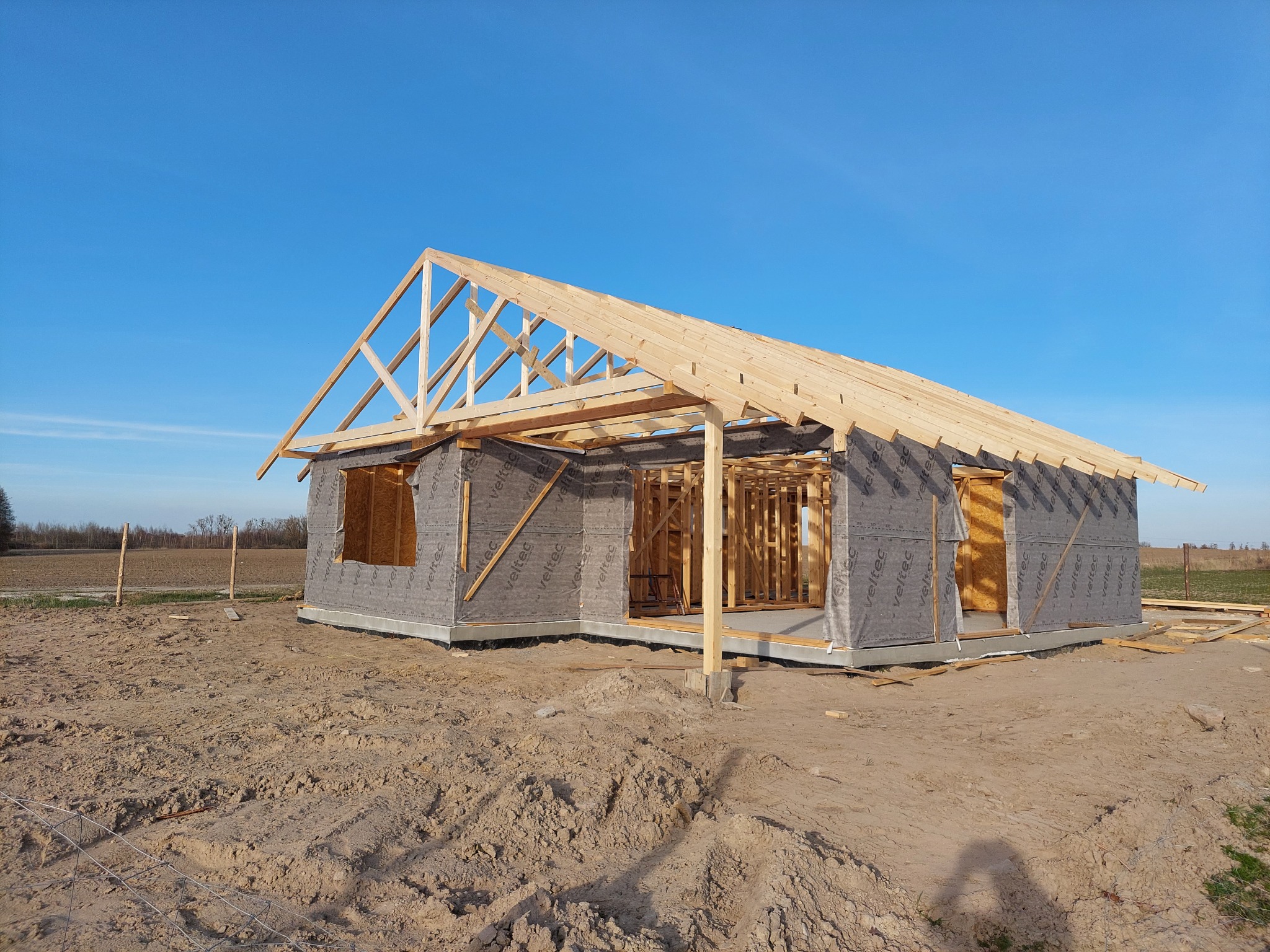 Surowy stan budowy domu: widoczna drewniana konstrukcja szkieletowa z zamontowaną membraną, odsłonięte krokwie dachu, teren budowy z piaskiem i błękitne niebo w tle.
