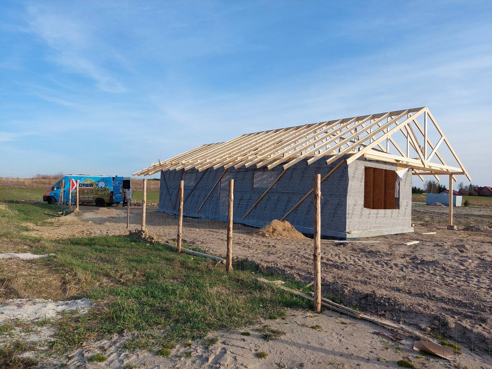 Budowa domu w technologii szkieletowej w trakcie montażu więźby dachowej, widoczny częściowo ocieplony budynek z szarą membraną, na tle błękitnego nieba i firmowy bus z reklamą.