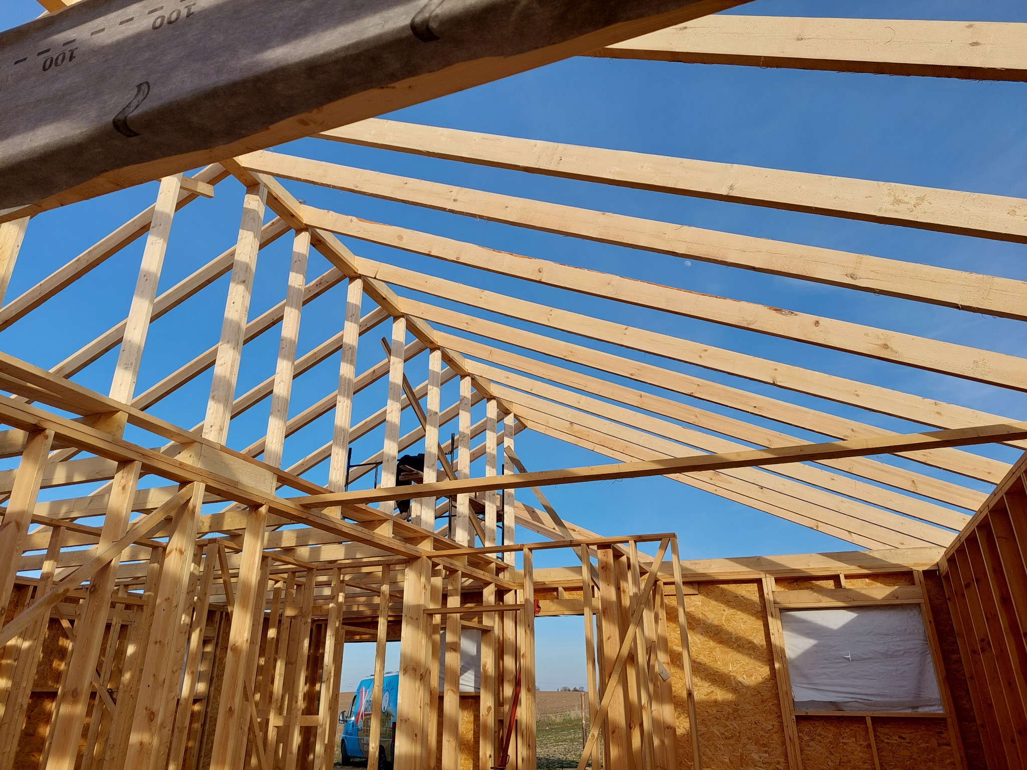 Wnętrze w trakcie budowy domu z drewnianą konstrukcją szkieletową, widoczne belki stropowe i ściany z płyt OSB, błękitne niebo w tle, pracownik na rusztowaniu wewnątrz konstrukcji.