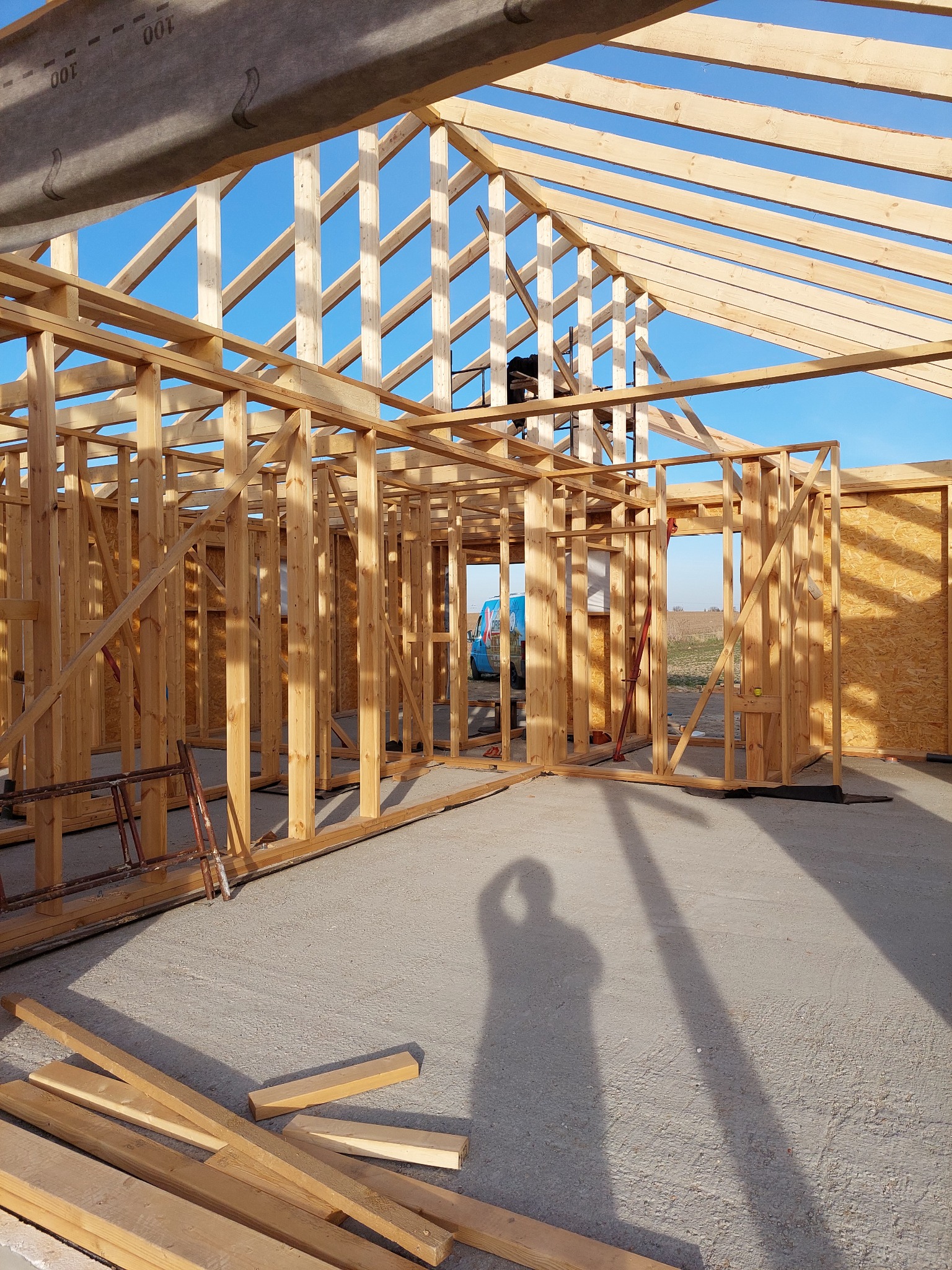 Konstrukcja domu w trakcie budowy, widoczne drewniane szkielety ścian i dachu na tle błękitnego nieba, cień osoby fotografującej na betonowej posadzce, rozrzucone fragmenty drewna.