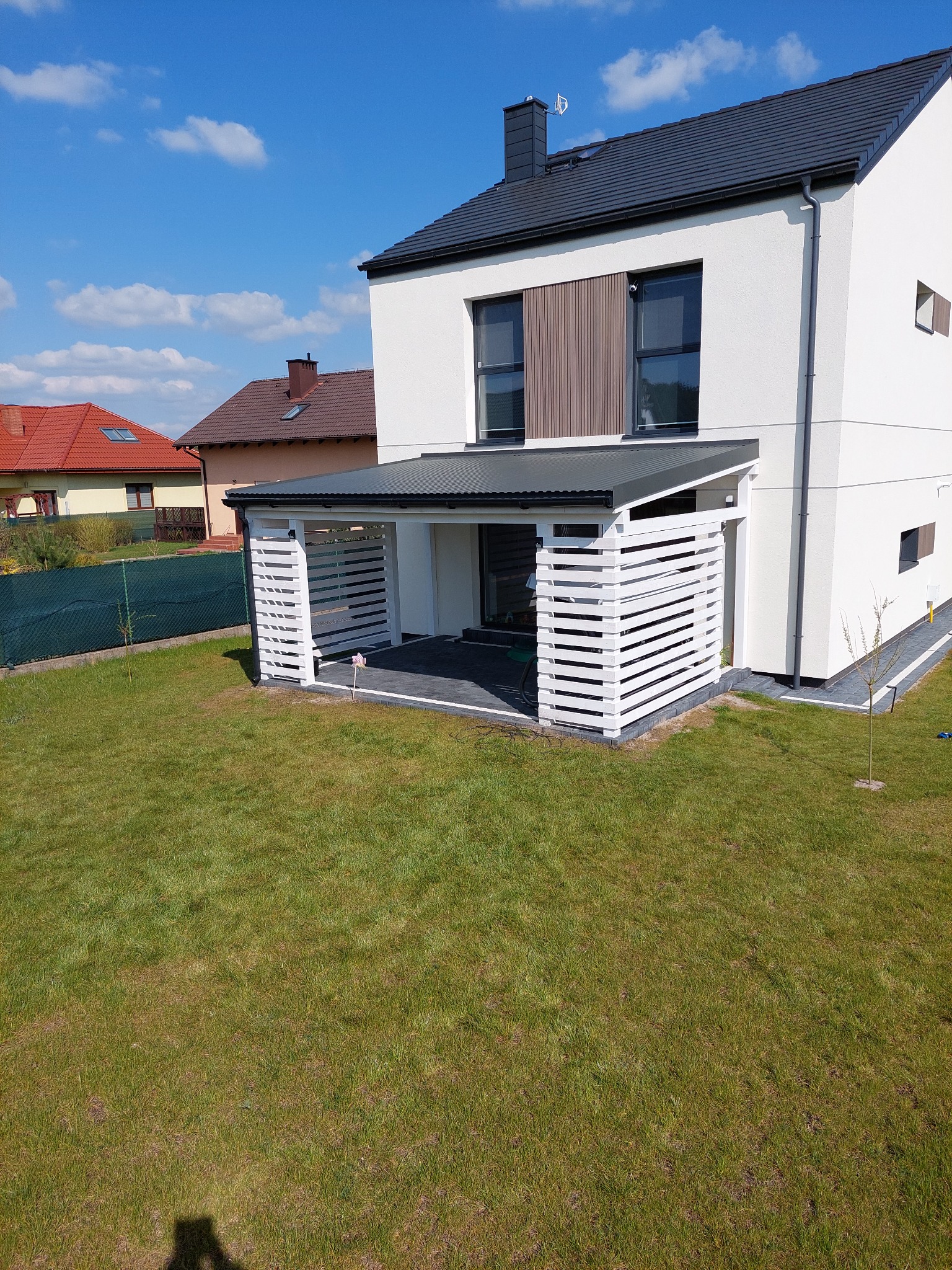 Nowoczesny dom z białymi ścianami i szarym dachem, z dobudowaną zadaszoną konstrukcją z białych, poziomych desek, tworzącą zacienioną przestrzeń tarasową z szarą podłogą, widok z trawnika...