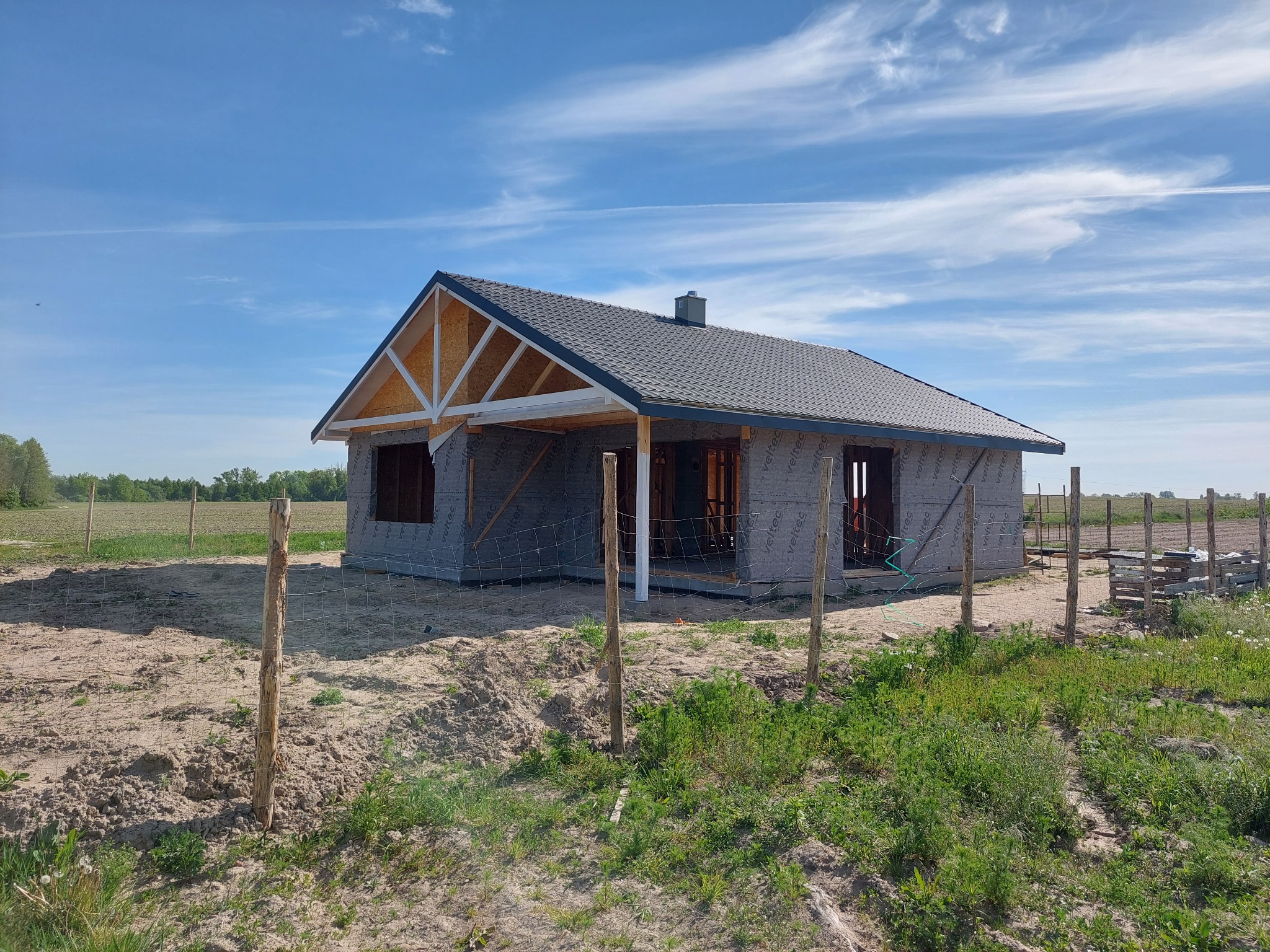 Niedokończony parterowy budynek z szarą dachówką i widoczną konstrukcją szkieletową dachu, zabezpieczony siatką ogrodzeniową, na piaszczystej działce z młodą zielenią, w tle pola uprawne...