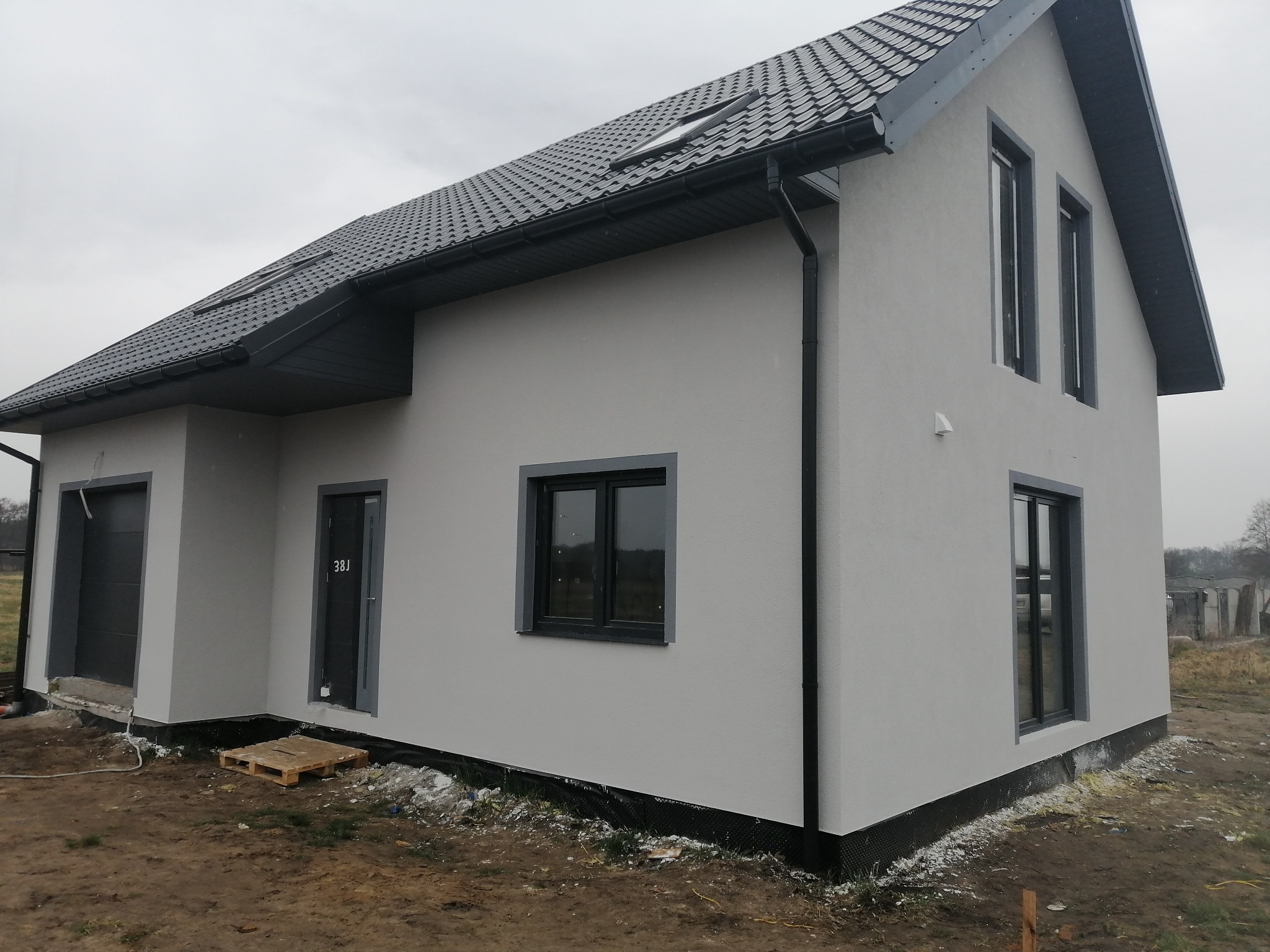 Nowoczesny dom szkieletowy z szarą elewacją i grafitową stolarką okienną, widoczne okna dachowe i paleta przy wejściu.