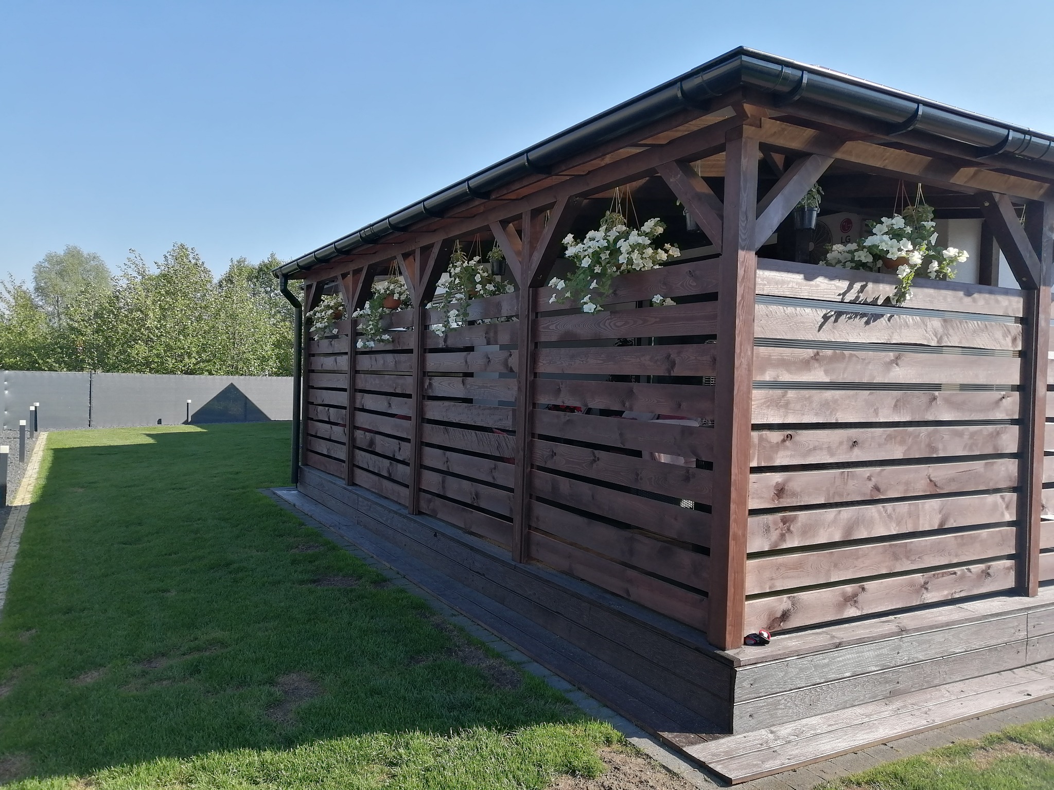 Zadaszona konstrukcja z ciemnego drewna z poziomymi panelami, ozdobiona wiszącymi doniczkami z białymi kwiatami, na tle zielonego trawnika i błękitnego nieba.