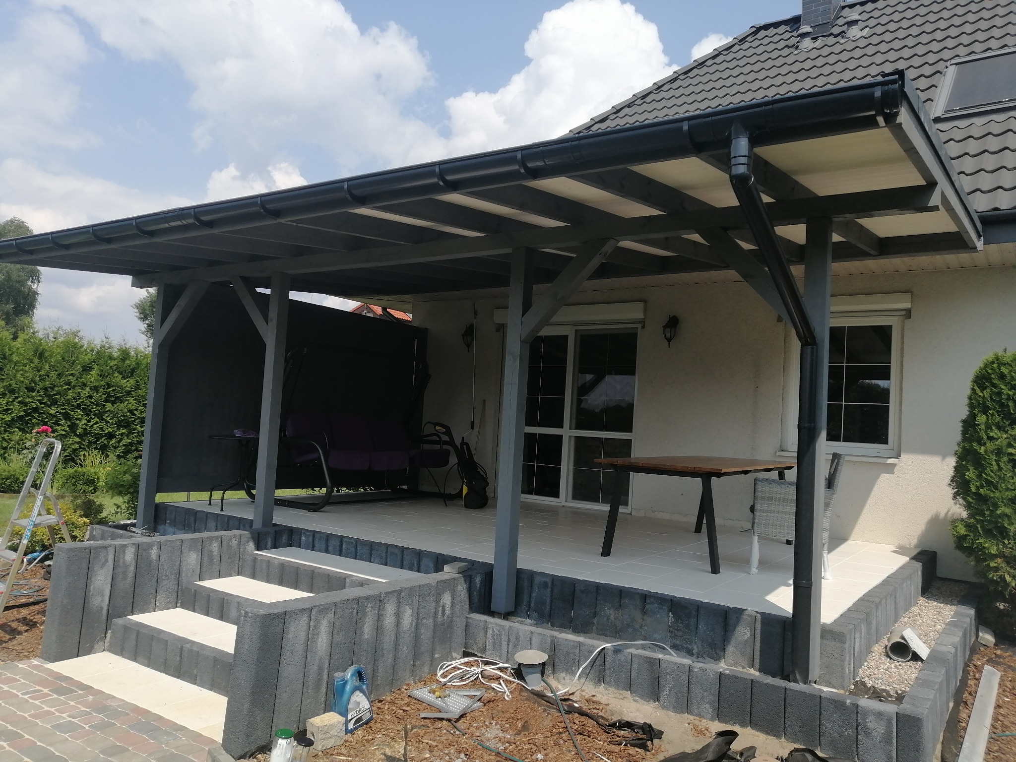 Zadaszony taras z drewnianą konstrukcją w ciemnoszarym kolorze, z widocznymi schodami z betonowych bloczków, meblami ogrodowymi i elementami wykończeniowymi w trakcie prac.