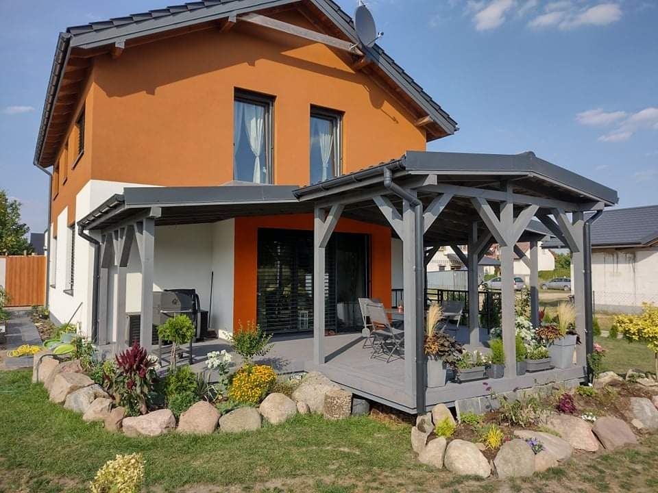 Nowoczesny dom z pomarańczową elewacją i szarą drewnianą pergolą połączoną z tarasem, otoczony ogrodem z kamieniami i roślinami.