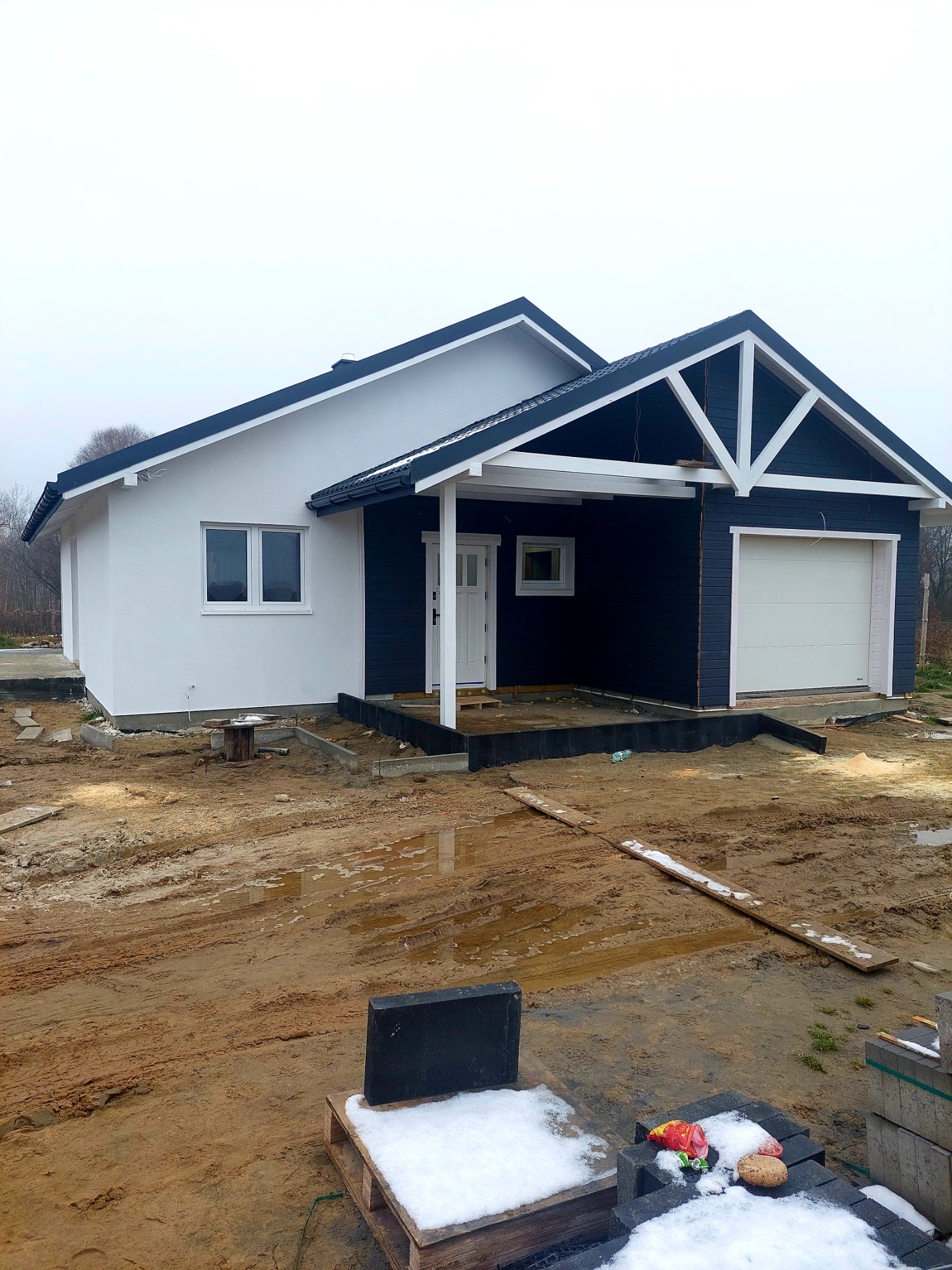 Niedokończony, parterowy dom z garażem, pomalowany na biało i granatowo, z widocznymi elementami konstrukcyjnymi z jasnego drewna, otoczony błotnistym terenem budowy z resztkami śniegu.