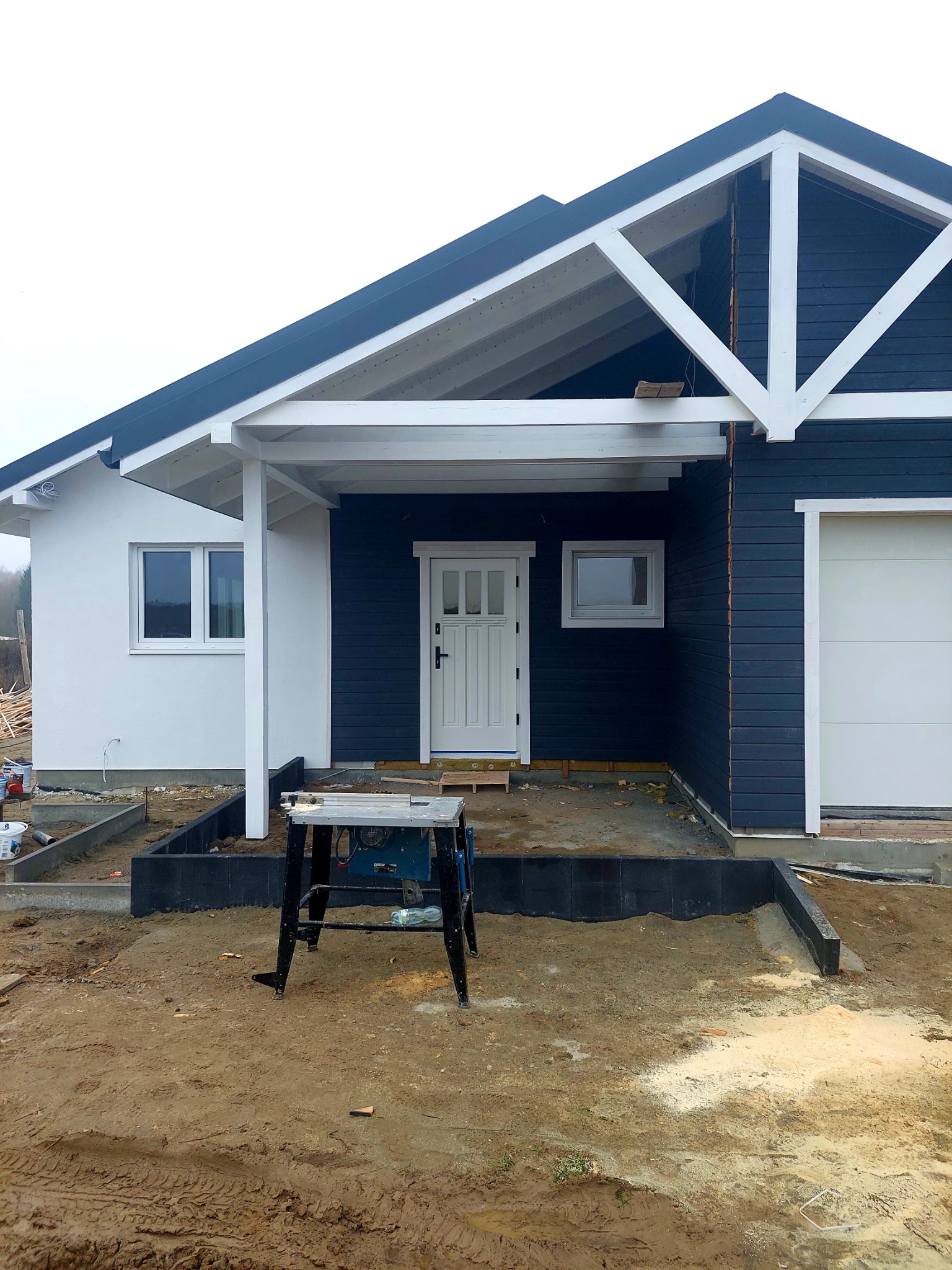 Niedokończony dom z ciemnego drewna z białymi detalami, widoczna piła stołowa na zewnątrz, teren wokół domu w trakcie prac ziemnych i przygotowania podłoża.