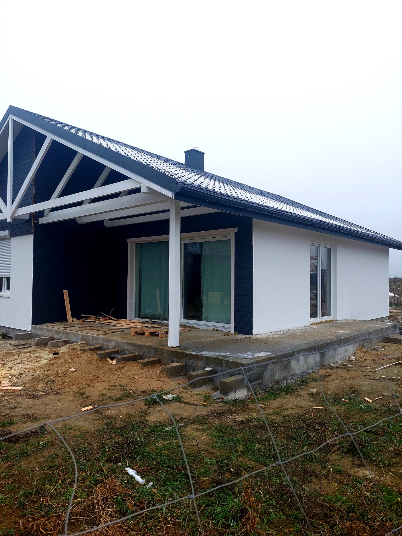 Niedokończony dom z czarno-białą elewacją i drewnianą konstrukcją dachu, widoczne materiały budowlane na tarasie i ziemi, ogrodzenie z siatki w pierwszym planie.