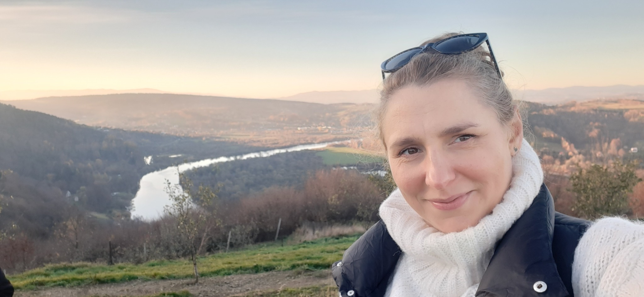 Autoportret kobiety w białym swetrze i czarnej kamizelce na tle rozległego krajobrazu z rzeką i wzgórzami o zachodzie słońca.