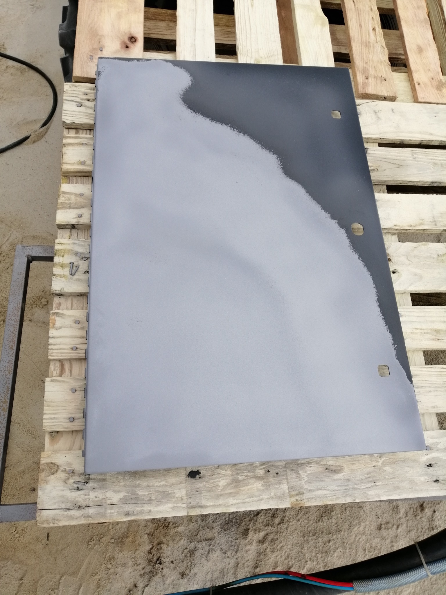 Metalowy arkusz częściowo pokryty szarą farbą, leżący na palecie z jasnego drewna. Widoczne otwory montażowe w górnej części arkusza.