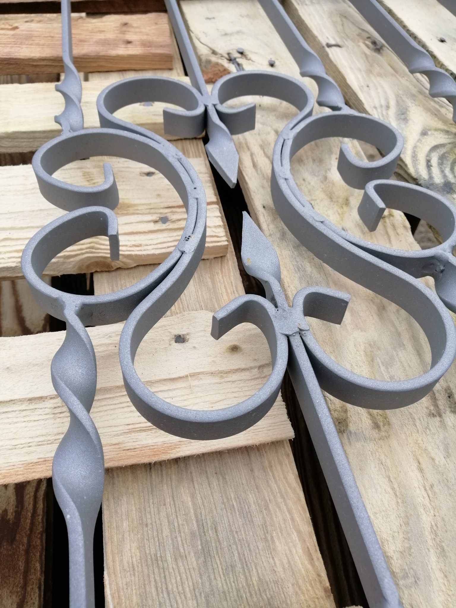 Dekoracyjne elementy metalowe z ornamentami, świeżo po obróbce, leżące na drewnianej palecie. Widoczna szara, matowa powierzchnia.