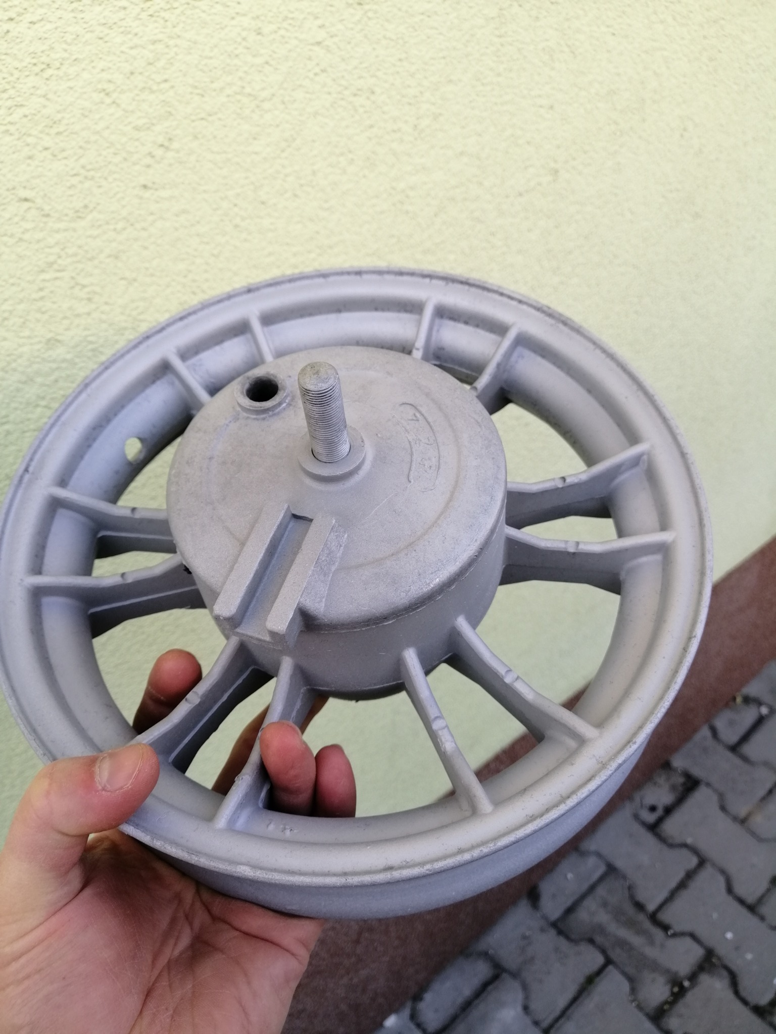 Szare, odnowione koło od skutera trzymane w dłoni na tle ściany i kostki brukowej.