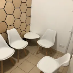 Poczekalnia z czterema białymi krzesłami, małym stolikiem i wieszakiem na ubrania, ściana z dekoracją w kształcie plastra miodu.