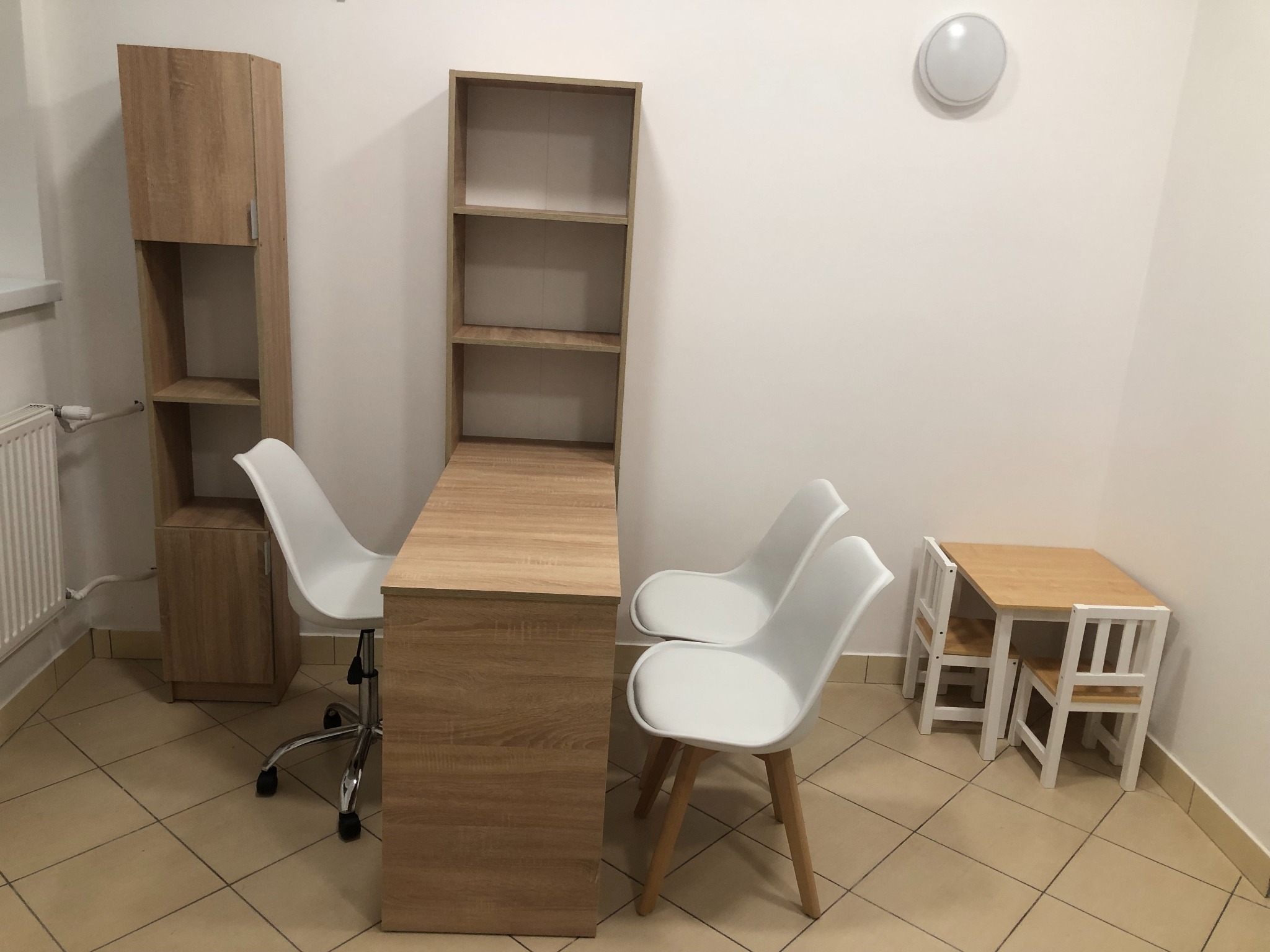 Gabinet z biurkiem i krzesłami w jasnych barwach, z drewnianymi elementami i białymi krzesłami, dodatkowo mały stolik z krzesełkami dla dzieci.
