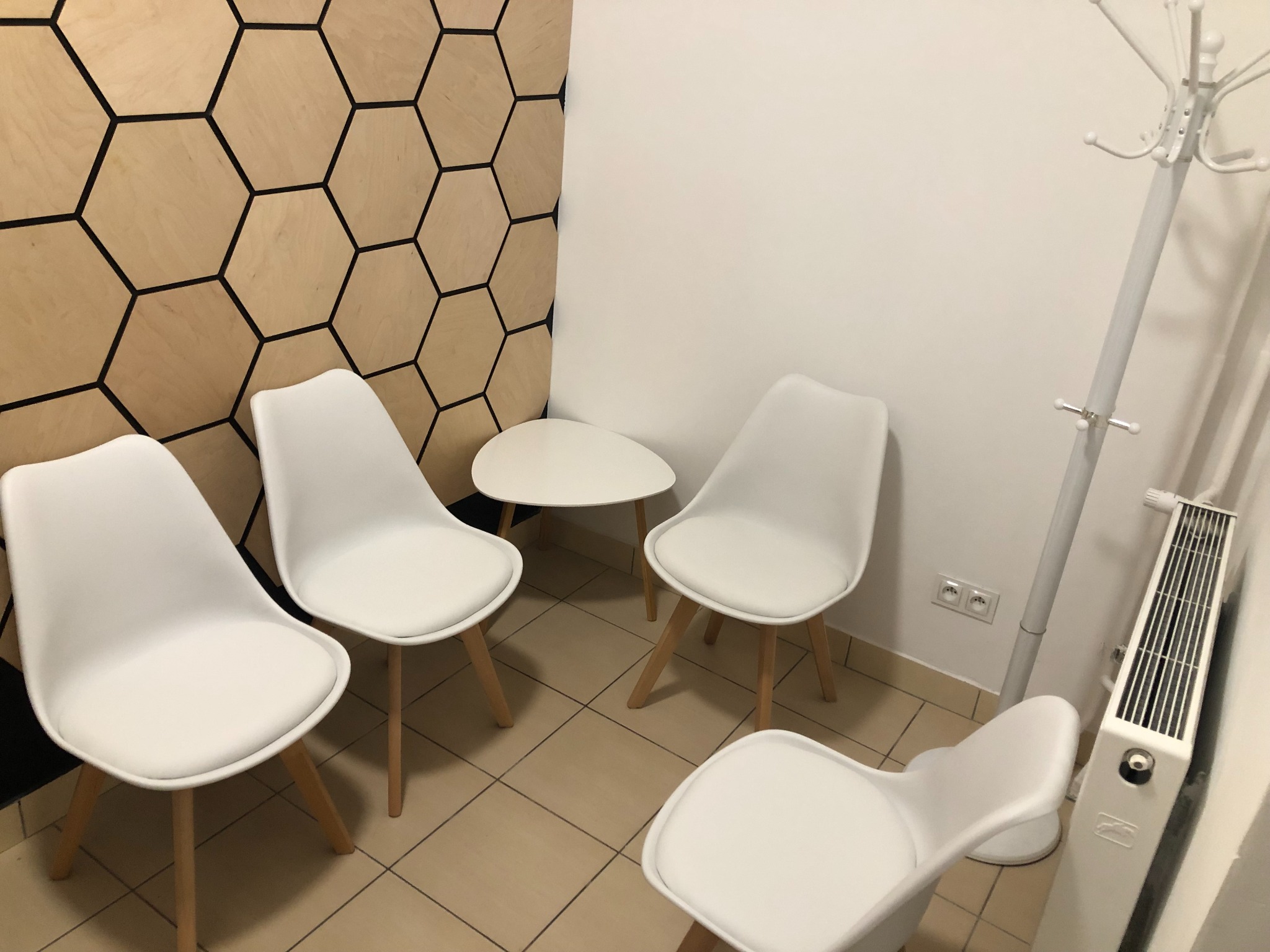 Poczekalnia z czterema białymi krzesłami, małym stolikiem i wieszakiem na ubrania, ściana z dekoracją w kształcie plastra miodu.