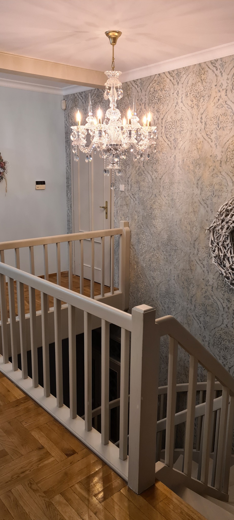 Kryształowy żyrandol oświetla klatkę schodową z drewnianą podłogą i balustradą, ściana w ozdobne wzory.
