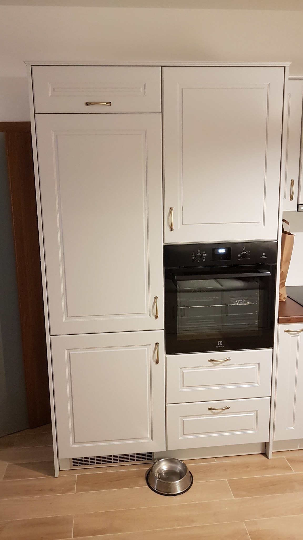 Kremowa zabudowa kuchenna z piekarnikiem Electrolux i złotymi uchwytami; na podłodze miska dla zwierząt.