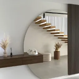Nowoczesne wnętrze z drewnianymi schodami wspornikowymi odbijającymi się w okrągłym lustrze, wisząca szafka z dekoracjami, oraz dekoracyjna ściana z pionowych paneli.