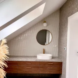 Nowoczesna łazienka na poddaszu z drewnianą szafką pod umywalką, okrągłym lustrem w złotej ramie i beżowymi płytkami na ścianie, uzupełniona dekoracyjną trawą pampasową.