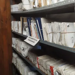 Metalowy regał wypełniony po brzegi segregatorami i plikami dokumentów, niektóre spięte sznurkiem, w archiwum firmy w Silnowie. Na jednej z półek widoczna kartka z napisem 'DOKUMENTACJA KSIĘGOWOŚĆ'.