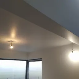 Pomieszczenie po remoncie, świeżo pomalowane ściany i sufit w jasnych odcieniach, duże okno z widokiem na zewnątrz w porannej mgle, dwie wiszące żarówki bez kloszy.