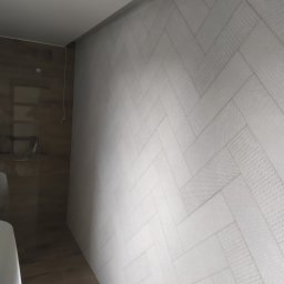 Fliz - Mal Jacek Bobrowski - Ściana w łazience wyłożona białymi płytkami ułożonymi we wzór jodełki, z delikatnym wzorem na każdej płytce, oraz fragment brązowej ściany z widocznymi przewodami.