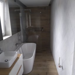 Fliz - Mal Jacek Bobrowski - Nowoczesna łazienka w Bęczarce z wolnostojącą białą wanną, umywalką nablatową na szafce z drewnianymi akcentami i drewnopodobną podłogą, ściana w geometryczny wzór, widoczne niedokończone elementy...