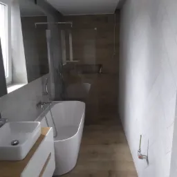 Nowoczesna łazienka w Bęczarce z wolnostojącą białą wanną, umywalką nablatową na szafce z drewnianymi akcentami i drewnopodobną podłogą, ściana w geometryczny wzór, widoczne niedokończone elementy...