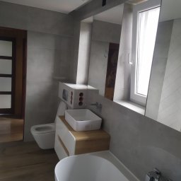 Fliz - Mal Jacek Bobrowski - Nowoczesna łazienka z szarą glazurą, drewnianą podłogą i białymi sanitariatami, odbicie lustrzane ukazuje fragment dekoracji ściennej.